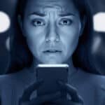 A woman with a concerned expression looks at her smartphone in a dimly lit environment, highlighting the impact of technology on emotions.