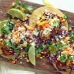 Three delicious tacos topped with fresh cilantro, diced tomatoes, and crumbled cheese, served on a wooden platter with lime wedges.