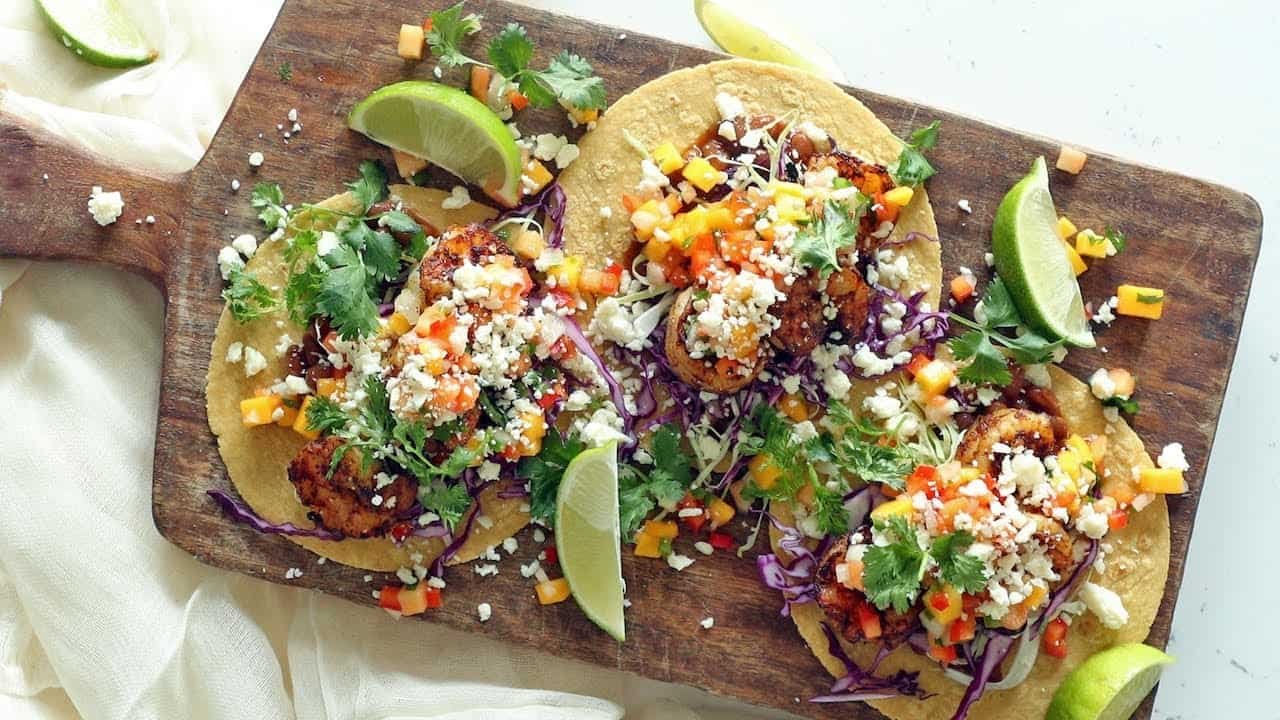 Three delicious tacos topped with fresh cilantro, diced tomatoes, and crumbled cheese, served on a wooden platter with lime wedges.