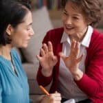 Two women engaged in a conversation, with one woman actively listening and taking notes, while the other gestures expressively, indicating a discussion about learning or mentorship.