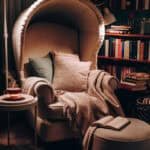 Cozy reading nook featuring a plush armchair with a high back, adorned with soft pillows and a warm blanket, beside a modern side table. In the background, a well-stocked bookshelf adds to the inviting atmosphere.