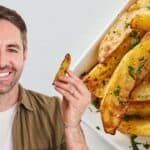 Smiling man holding a crispy potato wedge in front of a plate of golden-brown potato wedges garnished with herbs.