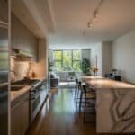 Modern kitchen interior featuring sleek cabinetry, a spacious island with unique marble design, and large windows allowing natural light. The open layout connects to a cozy living area with greenery, enhancing the inviting atmosphere.