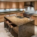 Modern kitchen interior featuring a spacious island with a granite countertop, stainless steel sink, and bar stools. The cabinetry is wood with a warm finish, complemented by under-cabinet lighting and a professional-grade stove, creating an inviting cooking space.