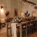 Elegant dining setup featuring a long wooden table adorned with a floral arrangement, surrounded by wooden chairs in a warmly lit interior. Decorative elements include soft lighting and stylish decor, creating a cozy atmosphere ideal for gatherings or events.