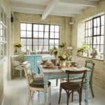 Bright and airy dining room featuring a rustic wooden table surrounded by various chairs, large windows with natural light, and floral centerpieces, creating a warm and inviting atmosphere.