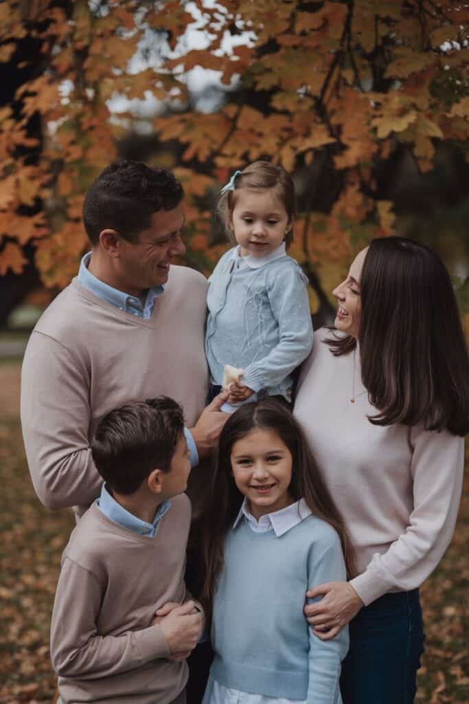 A happy family of five enjoying a fall day outdoors, surrounded by colorful autumn leaves. The father, wearing a light sweater, holds his young daughter, who is dressed in a blue cardigan. The mother, smiling, stands beside their two children, who are also in cozy sweaters. The scene captures a warm, joyful family moment in nature.