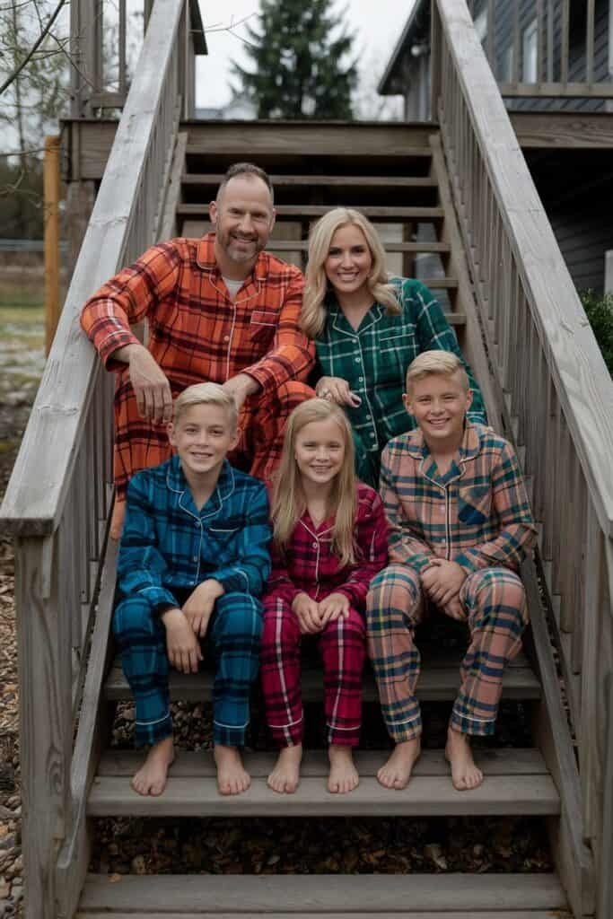 Family posing on wooden stairs wearing colorful matching pajamas. The group includes two adults and three children, showcasing a cozy, festive atmosphere.