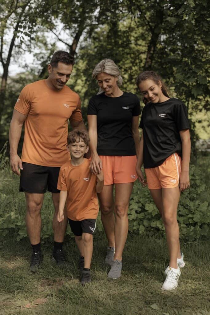 Family enjoying outdoor activities in matching athletic wear, featuring a father, mother, son, and daughter, surrounded by greenery. The adults wear orange and black shirts with shorts, while the children sport bright and playful outfits. Ideal for promoting family fitness and active lifestyle.