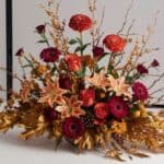 Vibrant autumn floral arrangement featuring red and orange flowers, including lilies, gerbera daisies, and decorative branches, set against a neutral background. Perfect for seasonal decor or special occasions.