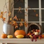 Autumn-themed decorative display featuring a variety of pumpkins, gourds, and fall foliage in a vase, arranged on a table near a window.