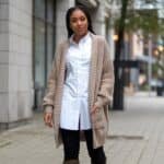 Young woman wearing a long beige cardigan over a white shirt and black leggings, walking on a city sidewalk with trees and buildings in the background.