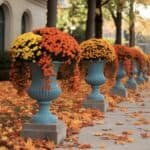 Colorful autumn chrysanthemums in blue urns line a pathway, surrounded by fallen leaves, creating a vibrant seasonal display.