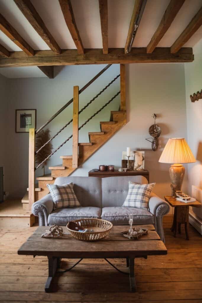 Cozy living room interior featuring a gray sofa with plaid cushions, a wooden coffee table with decorative items, and a staircase in the background. Soft lighting from a lamp creates a warm ambiance.