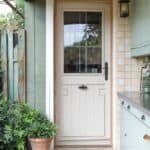 Exterior view of a cream-colored door with a window, surrounded by greenery and a patio area, showcasing a modern entryway design.