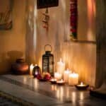 Cozy interior scene featuring a warm glow from multiple candles and a lantern, set against a textured wall adorned with traditional decor and flowers, creating a serene ambiance.