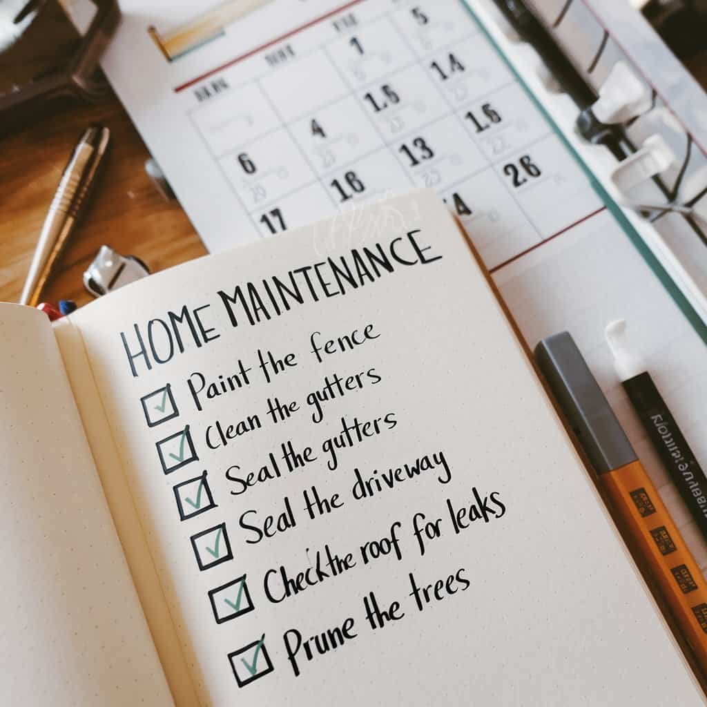 Image of a handwritten home maintenance checklist on a notebook, featuring tasks like painting the fence, cleaning gutters, sealing the driveway, checking roof for leaks, and pruning trees, with a calendar and pens in the background.