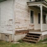 Weathered exterior of a house showing peeling paint on wood siding and worn wooden steps leading to the porch, highlighting the need for renovation and maintenance.