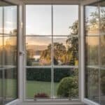 View of a tranquil garden landscape through a large window, showcasing lush greenery and a sunset sky in the background.