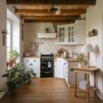 Cozy kitchen interior featuring wooden beams, white cabinetry, and a black stove. Natural light illuminates potted plants and a wooden table, creating a warm and inviting atmosphere.