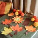 Colorful autumn-themed paper crafts displayed on a green table, featuring smiling sun and leaf shapes in orange and yellow, complemented by decorative pom-poms and a wooden "Hello Fall" sign.