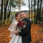 Couple in formal wedding attire posing in a forest during autumn, surrounded by colorful leaves, with the bride holding a bouquet of red flowers.