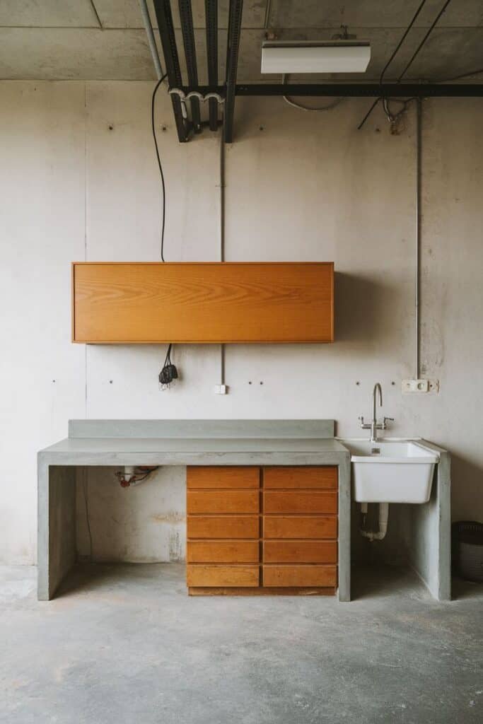 Modern industrial kitchen workspace featuring a concrete countertop, wooden drawers, and a white sink. The minimalist design includes an overhead wooden cabinet and exposed ceiling elements, showcasing a blend of functionality and contemporary style.