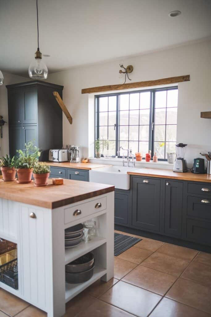 Modern kitchen interior featuring a blend of dark green cabinetry and a white kitchen island. The space includes wooden countertops, a farmhouse sink, and stylish appliances. Natural light filters through a large window adorned with potted plants, enhancing the inviting atmosphere.