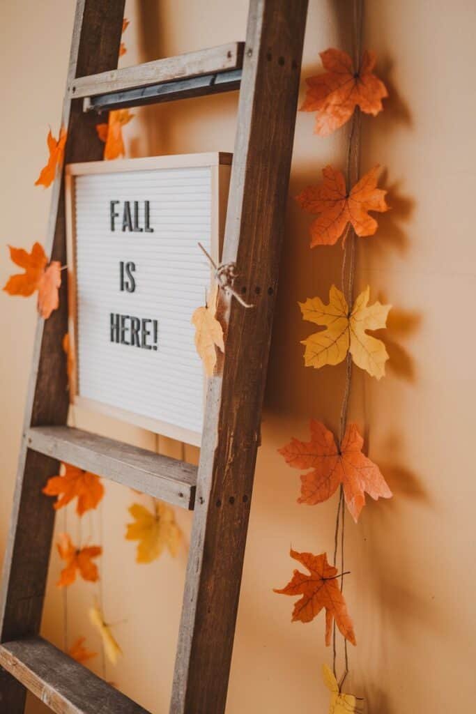 Wooden ladder decorated with colorful artificial fall leaves and a sign that reads "FALL IS HERE!" against a warm beige wall, creating a cozy autumn atmosphere.