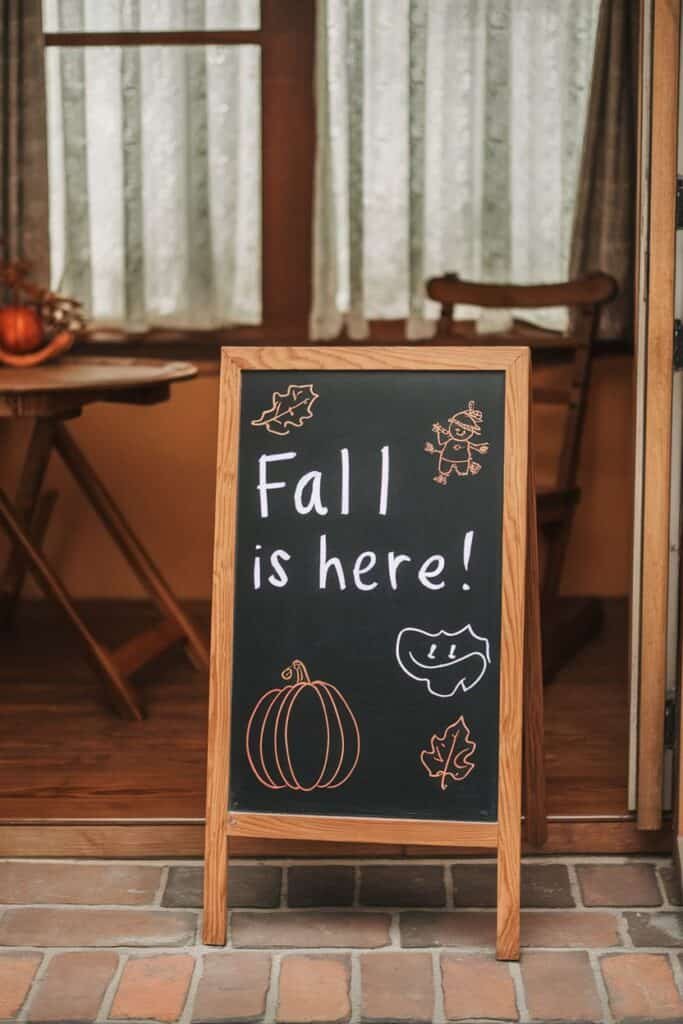 Chalkboard sign announcing "Fall is here!" with autumn-themed illustrations, including a pumpkin and leaves, displayed outside a cozy cafe with wooden furniture and warm interior lighting.