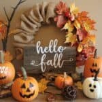 Autumn-themed decor featuring a burlap wreath adorned with colorful leaves, a wooden sign that reads "hello fall," and various pumpkins, including carved jack-o'-lanterns and decorative white pumpkins, set against a warm background.