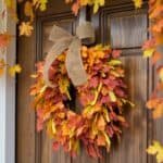 Autumn-themed wreath made of vibrant orange, red, and yellow leaves, hanging on a wooden door, adorned with a burlap bow, creating a seasonal decor accent.