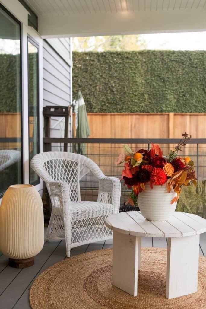 Cozy outdoor patio featuring a white wicker chair, a round wooden table, and a decorative vase filled with vibrant fall flowers. A soft rug adds warmth to the space, complemented by a stylish lamp, creating an inviting atmosphere for relaxation.