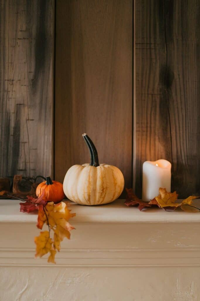 Autumn-themed decor featuring a white pumpkin, a small orange pumpkin, colorful fall leaves, and a lit candle on a mantel against a rustic wooden background.