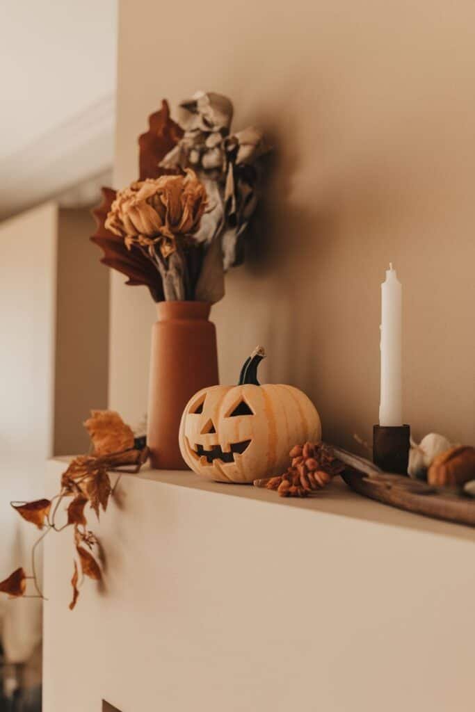 Autumn-themed mantel decor featuring a carved pumpkin, dried flowers in a terracotta vase, a white candle, and decorative leaves, creating a cozy seasonal atmosphere.