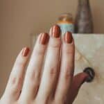 Close-up of a hand with neatly manicured, rust-colored nails, set against a softly lit background featuring decorative items.