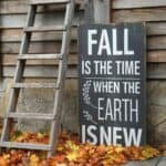 Wooden sign with autumn-themed quote about fall, surrounded by colorful leaves and a rustic ladder against a weathered wooden wall.