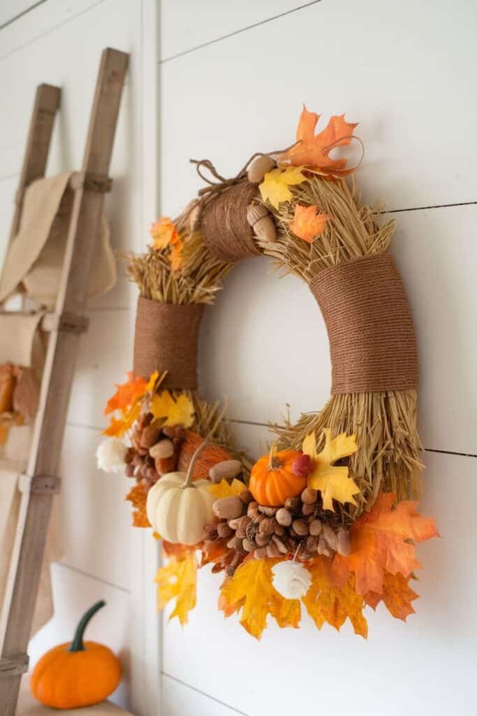 Autumn-themed wreath decorated with straw, colorful leaves, acorns, and miniature pumpkins, hanging on a white wall alongside a wooden ladder and small orange pumpkin. Perfect for seasonal home decor.