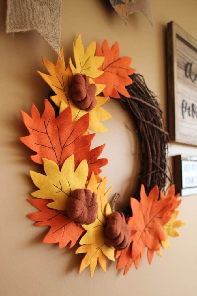 Colorful felt autumn wreath featuring orange, yellow, and brown leaves with small fabric pumpkins, displayed on a wall. Ideal for fall home decor and seasonal celebrations.