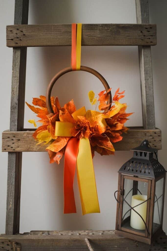 Autumn wreath made of vibrant orange and yellow leaves, adorned with a yellow and orange ribbon, hanging on a rustic wooden ladder next to a vintage lantern with a candle. Perfect decor for fall celebrations.