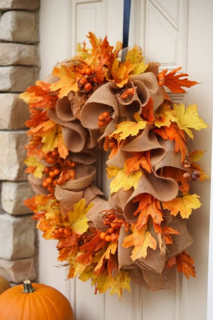 Autumn-themed wreath made of burlap, vibrant orange and yellow leaves, and decorative berries, hanging on a door with a stone wall background. Perfect for seasonal home decor.