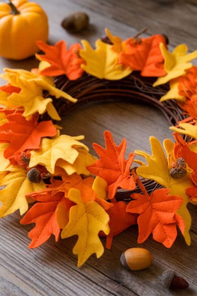 Colorful autumn wreath made of felt leaves in shades of orange, yellow, and red, surrounded by decorative pumpkins and acorns on a wooden surface, perfect for fall home decor.