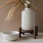 White ceramic vase with textured leaf patterns, holding dried pampas grass, paired with a matching bowl on a wooden stand, set against a neutral background. Ideal for home decor and interior styling.