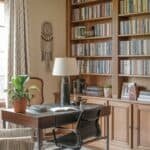 Cozy home office with a wooden desk, black chair, and a potted plant, featuring a large bookshelf filled with books, warm lighting, and decorative elements.