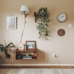 Cozy interior living space featuring a light-colored wall with a wall clock, framed artwork, a wooden shelf with a picture frame, and a potted plant. A lamp hangs above, and a trailing plant adds a touch of greenery to the decor.