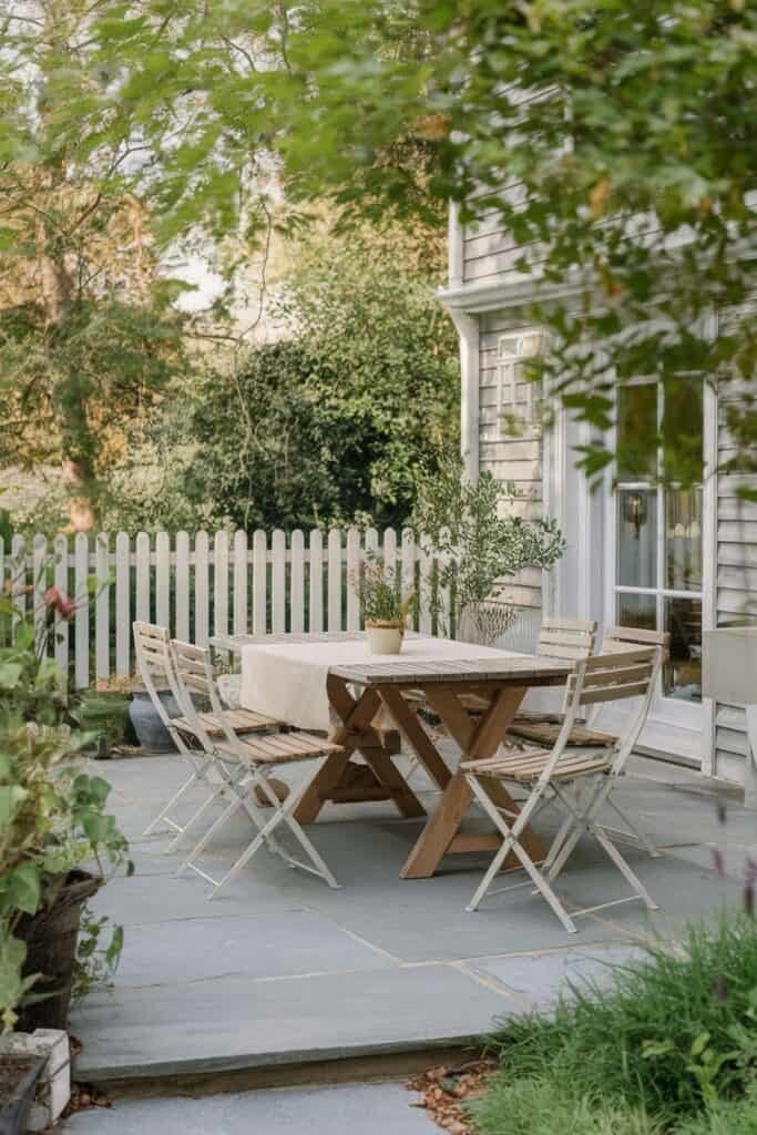 Outdoor dining area featuring a wooden table and white folding chairs, surrounded by greenery and a white picket fence, ideal for alfresco meals.