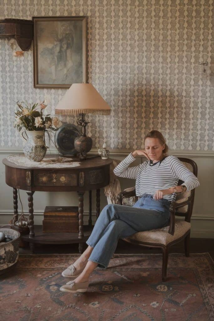 Woman sitting in a vintage chair, wearing a striped shirt and relaxed jeans, in a cozy, elegantly decorated room with patterned wallpaper, a decorative table, and a lamp. A vase of flowers and various antiques are visible, creating a warm and inviting atmosphere.